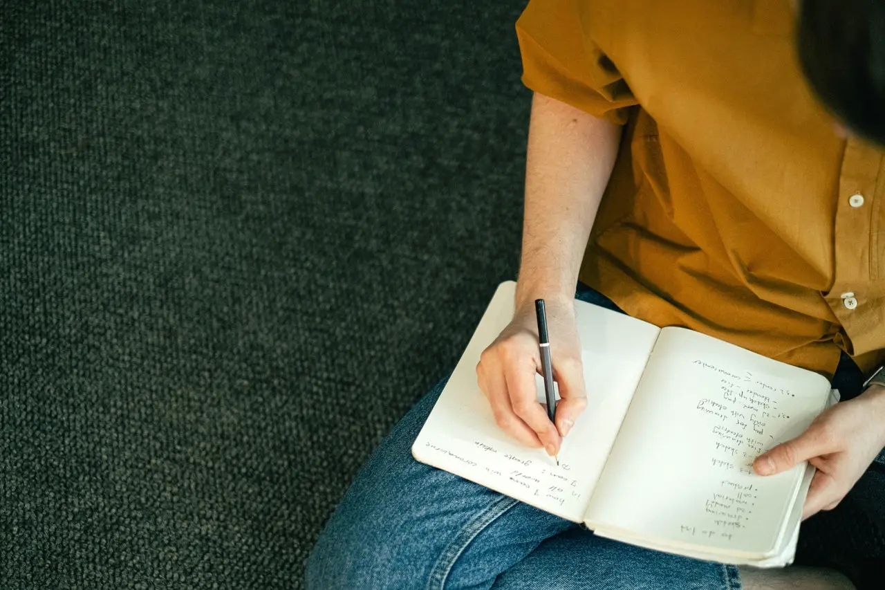 A person sits on the floor writing a handwritten checklist in a notebook, capturing the planning involved in a house move.