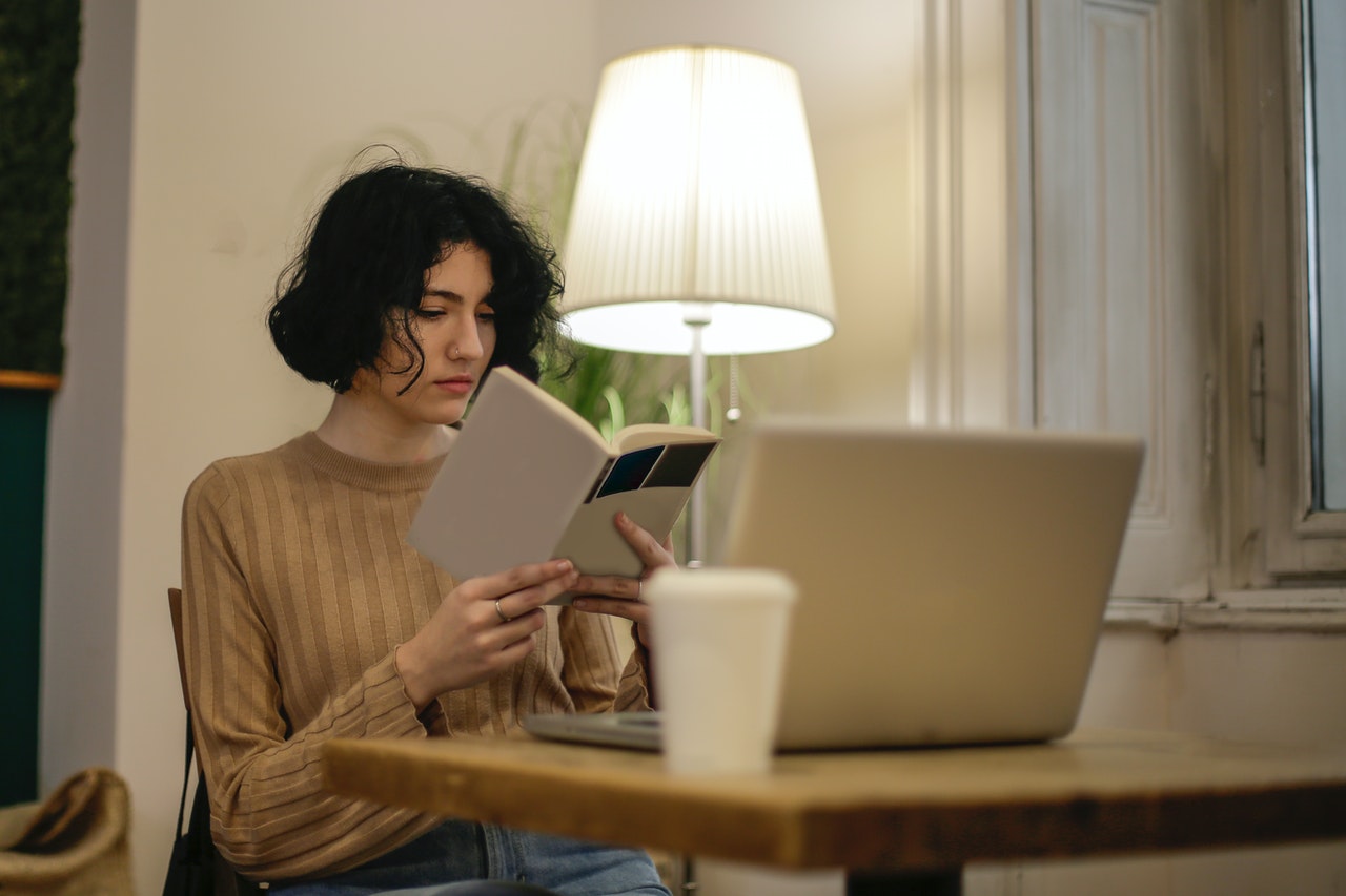 A young tenant reads a book by a table lamp, a laptop and coffee cup on table in front