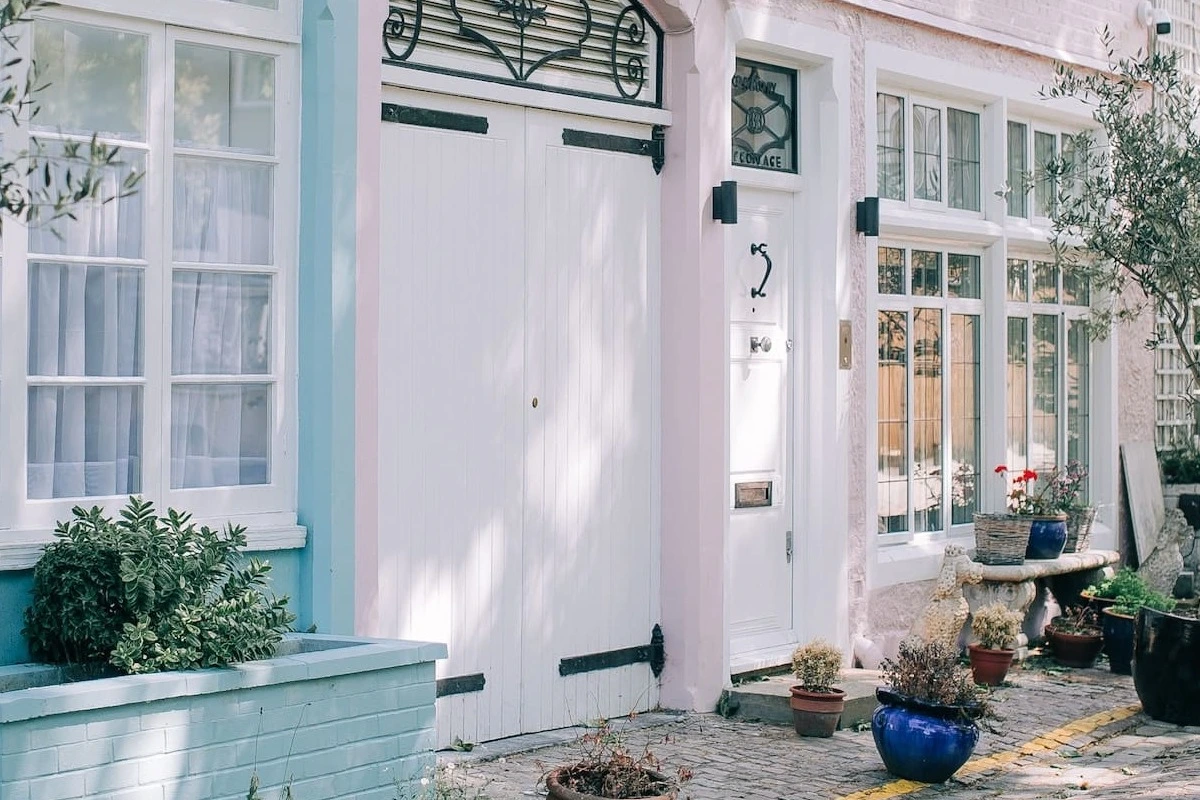 A pastel-painted London mews house frontage with white carriage doors, sash windows and potted plants on a cobbled lane.