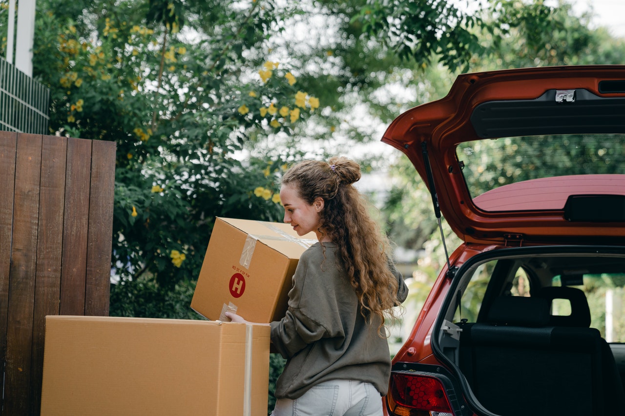Young woman carries a cardboard moving box from the boot of a red car to her new house, other boxes lie around 