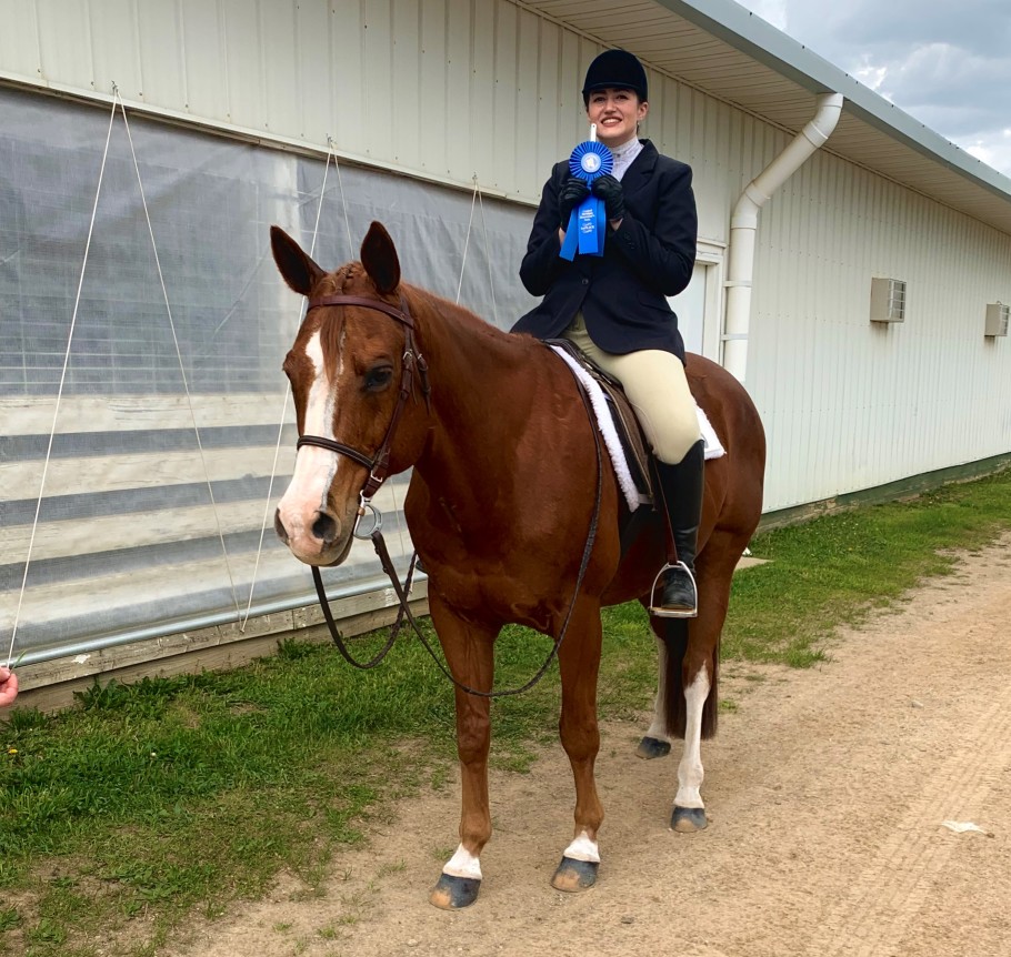 Woman in hunter show clothes holding first-place ribbon on chestnut horse w/ white blaze