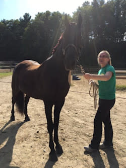 Woman smiling while holding dark horse in halter with woods and sunrays in background