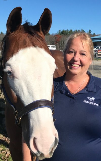 A sorrel Quarter Horse with a white face stands next to his human, Linda, who wears a navy blue polo shirt with a white SmartPak logo. 