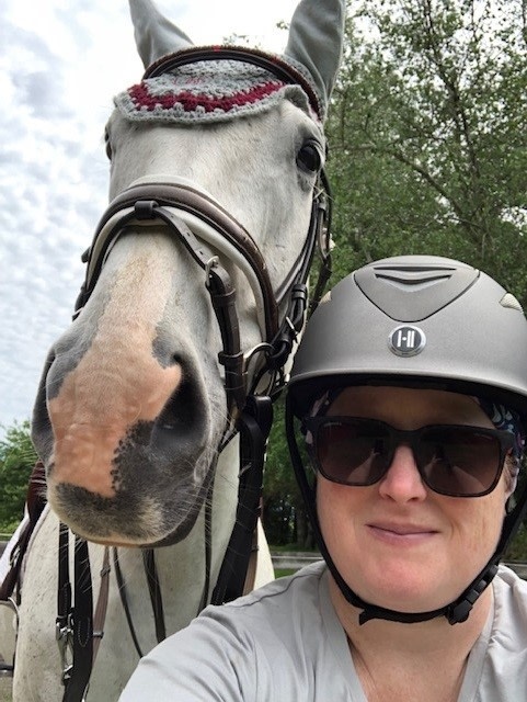 Selfie of woman in helmet with gray Thoroughbred wearing gray and red ear bonnet