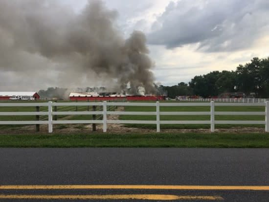 White fencing in front of a barn smoking on fire in the distance.