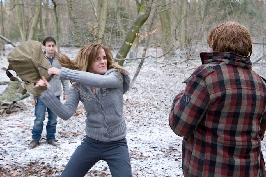 Hermione and Ron outside in a snowy wood. She is angrily hitting him with a bag and Harry is stood in the background.