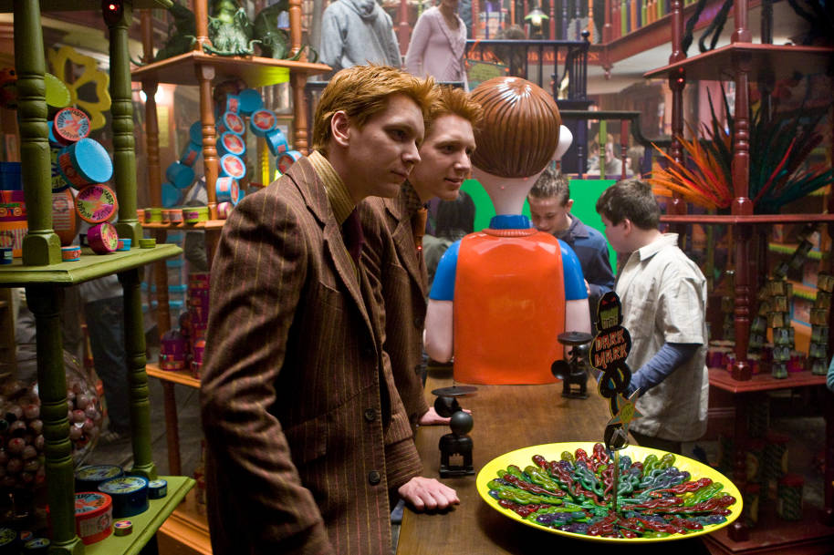 Fred and George Weasley standing behind a table in Weasleys' Wizard Wheezes while surrounded by products.