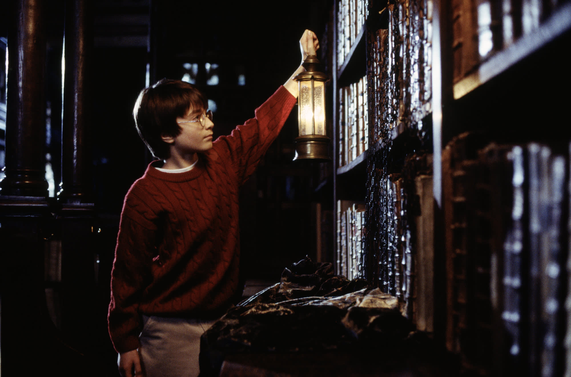 Harry holding a lantern up to look at the books in the Restricted Section of the Hogwarts Library. His Invisibility Cloak is close by. 