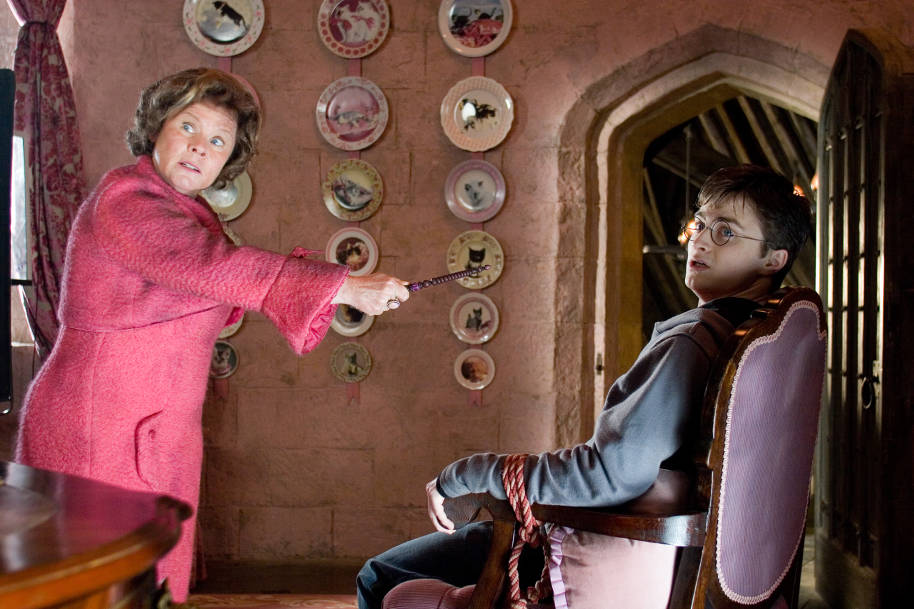 Harry is tied to a chair in Umbridge's office looking shocked. She is standing over him, pointing her wand in his face and looks dishevelled and out of control.