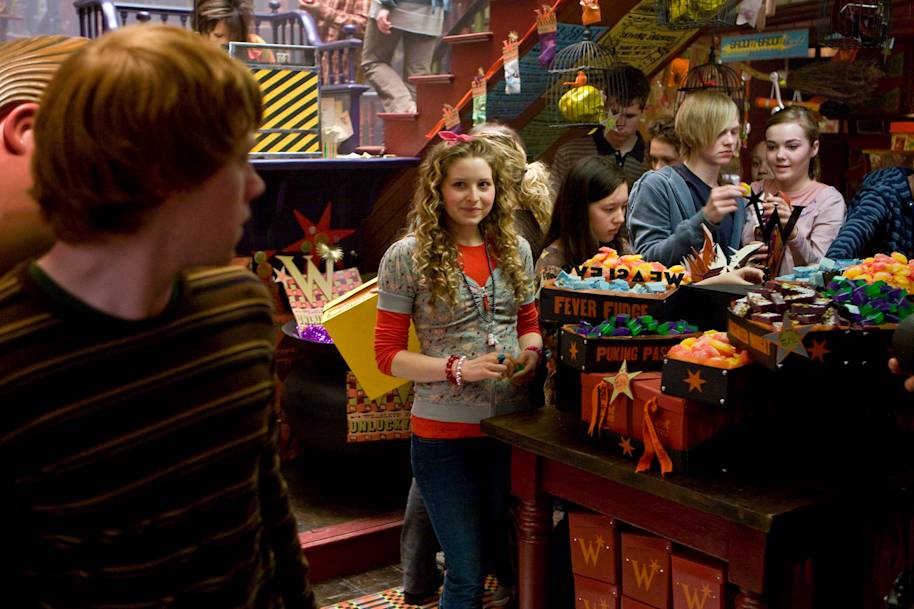 Inside Weasleys' Wizard Wheezes. Lavender Brown is standing by Skiving Snackbox products and smiling in Ron Weasley's direction.