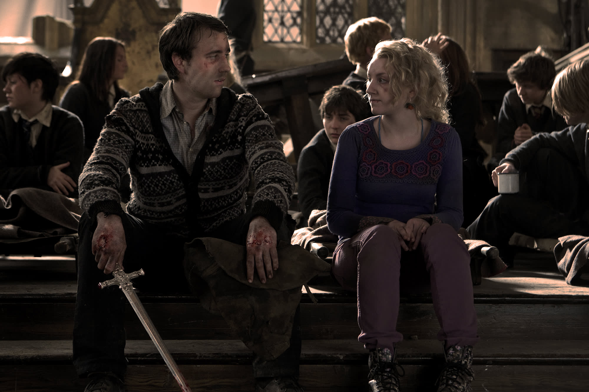 Luna and Neville sat on a table in the Great Hall after the Battle of Hogwarts. Both have cuts and bruises and Neville is holding the sword of Gryffindor. 