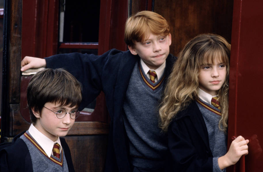 Harry, Ron and Hermione by the Hogwarts Express. Hermione and Ron are leaning out of one the doors while Harry is next to them.They are all smiling slightly. 
