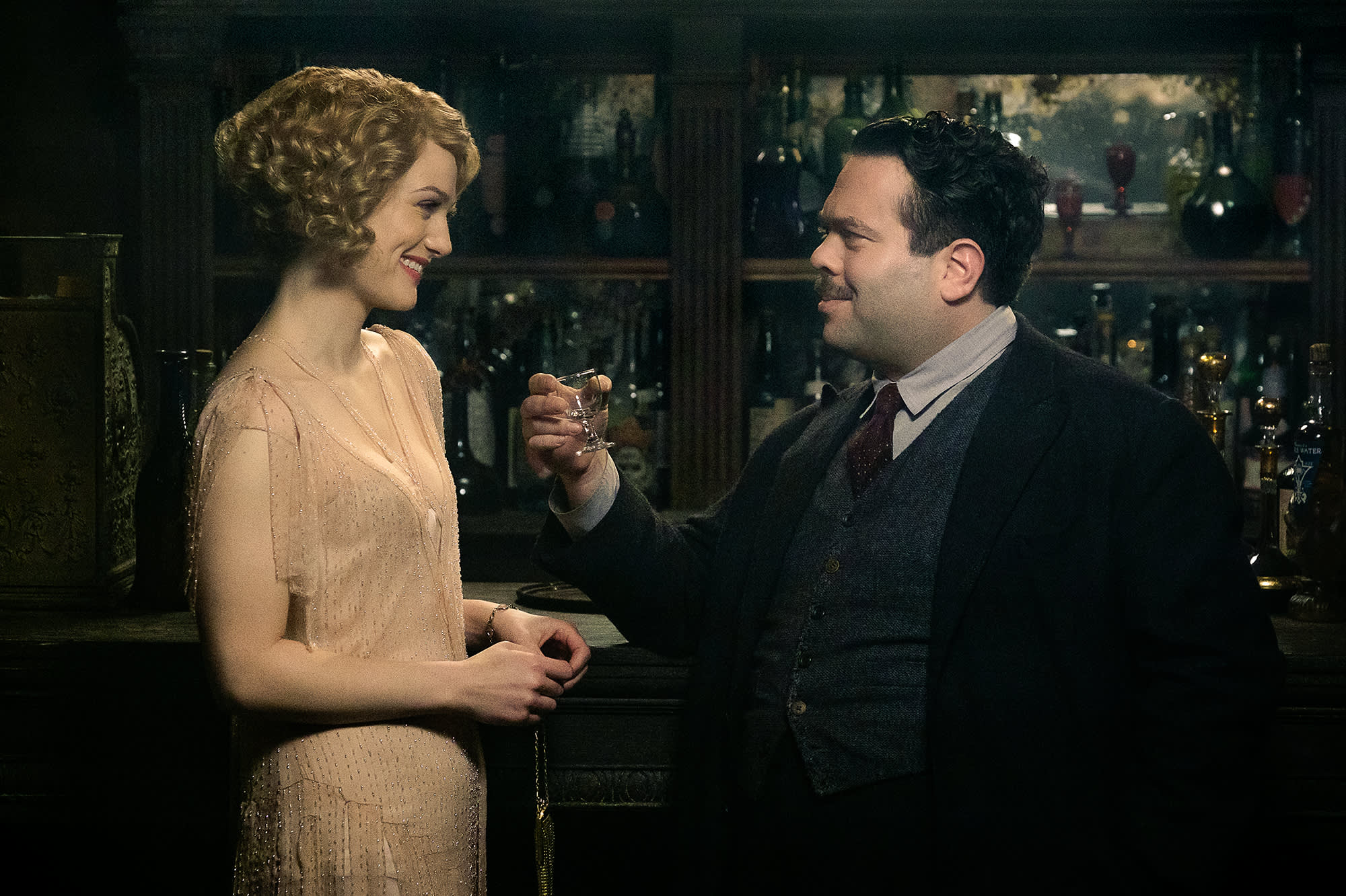 Jacob and Queenie standing at the bar of The Blind Pig looking at each other and smiling. Jacob is holding a glass of Gigglewater.