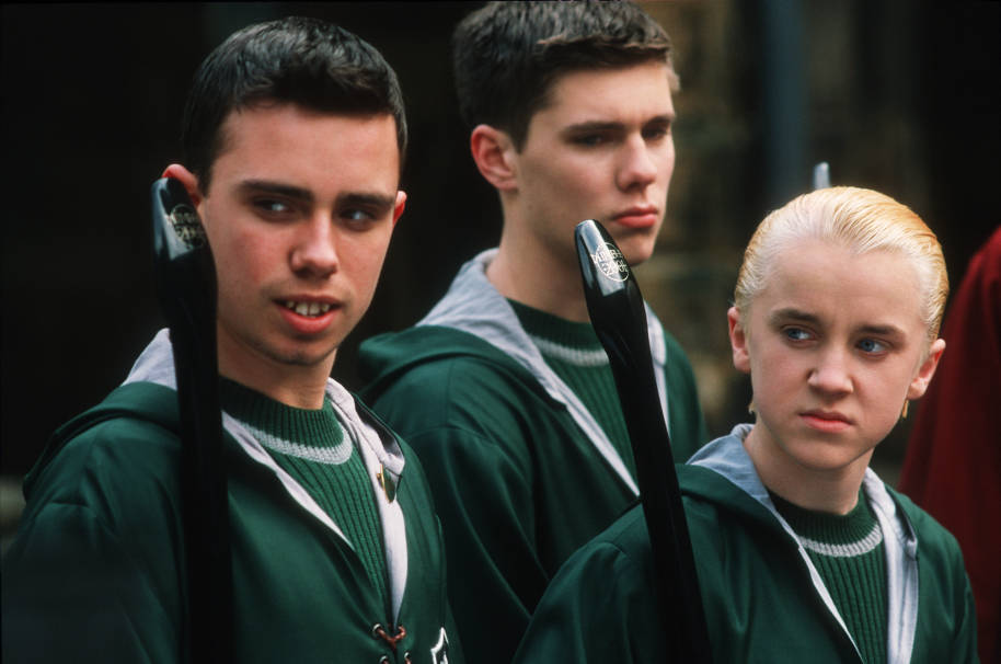 Draco Malfoy is standing with other members of the Slytherin Quidditch team. They are wearing Quidditch robes and holding their Nimbus 2001 broomsticks.