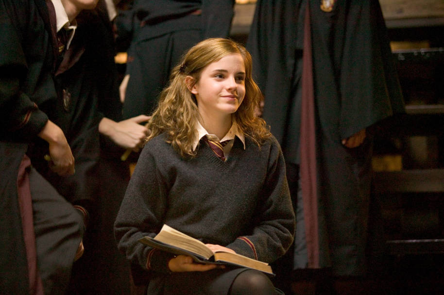Hermione sat on a bench in the Great Hall. She has a book open on her lap and is smiling.