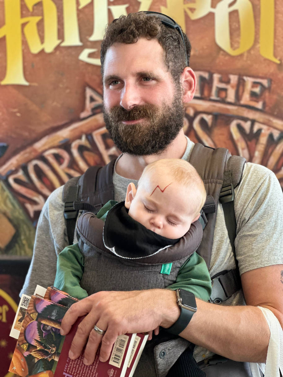A man carrying a sleeping baby in a baby carrier. The baby is dressed up as Harry Potter with his scar.