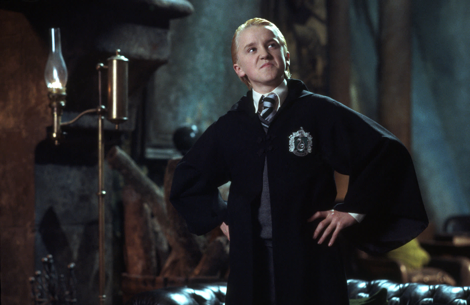 Draco Malfoy with his hands on his hips, looking defiant in the Slytherin Common Room of Hogwarts castle.