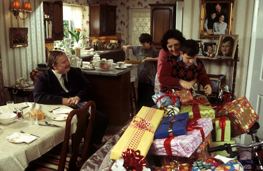 The Dursleys celebrating Dudley's birthday. Harry is cooking bacon in the kitchen, Vernon is sat at the table and Petunia is hugging Dudley who is surrounded by a huge pile of presents.