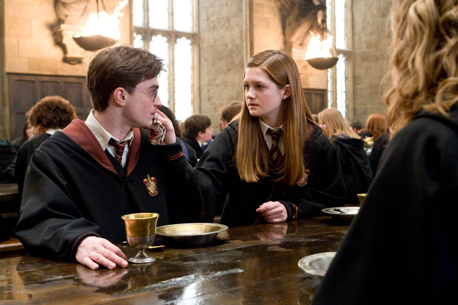 Ginny and Harry are sat in the Great Hall. Ginny is wiping Harry's mouth with a cloth.