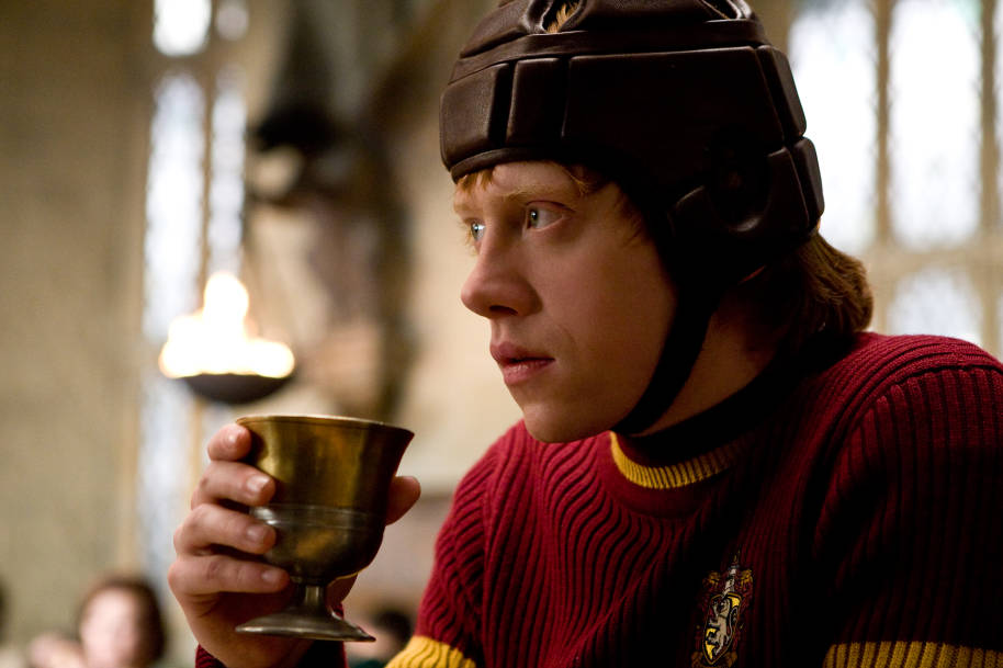Ron is sitting in the Great Hall in his Quidditch robes. He is holding a goblet about to take a drink. 