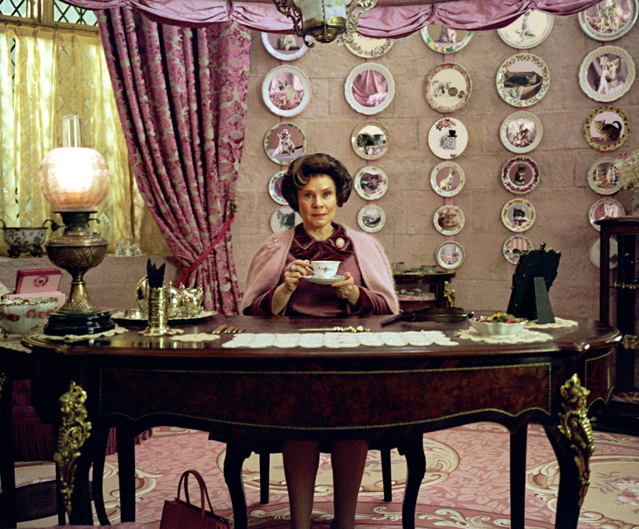Dolores Umbridge is sat at her desk in her office, drinking tea and looking smug. Her wall of cat plates in behind her.
