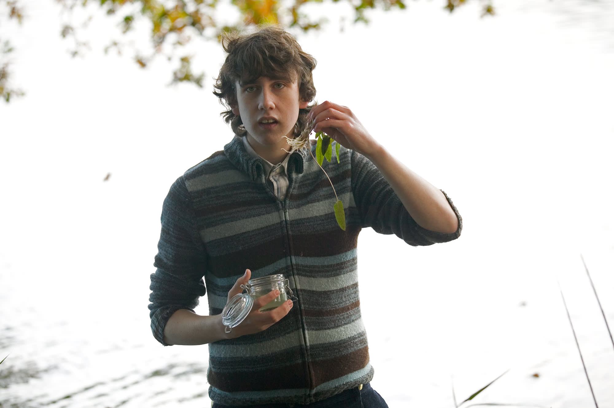 Neville Longbottom stands at the edge of the lake while holding up a plant