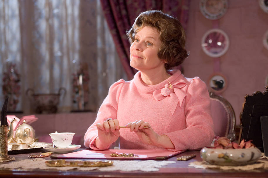 Umbridge is sat at her desk in her office and looking up at someone of shot. She is holding a pencil in her hands.