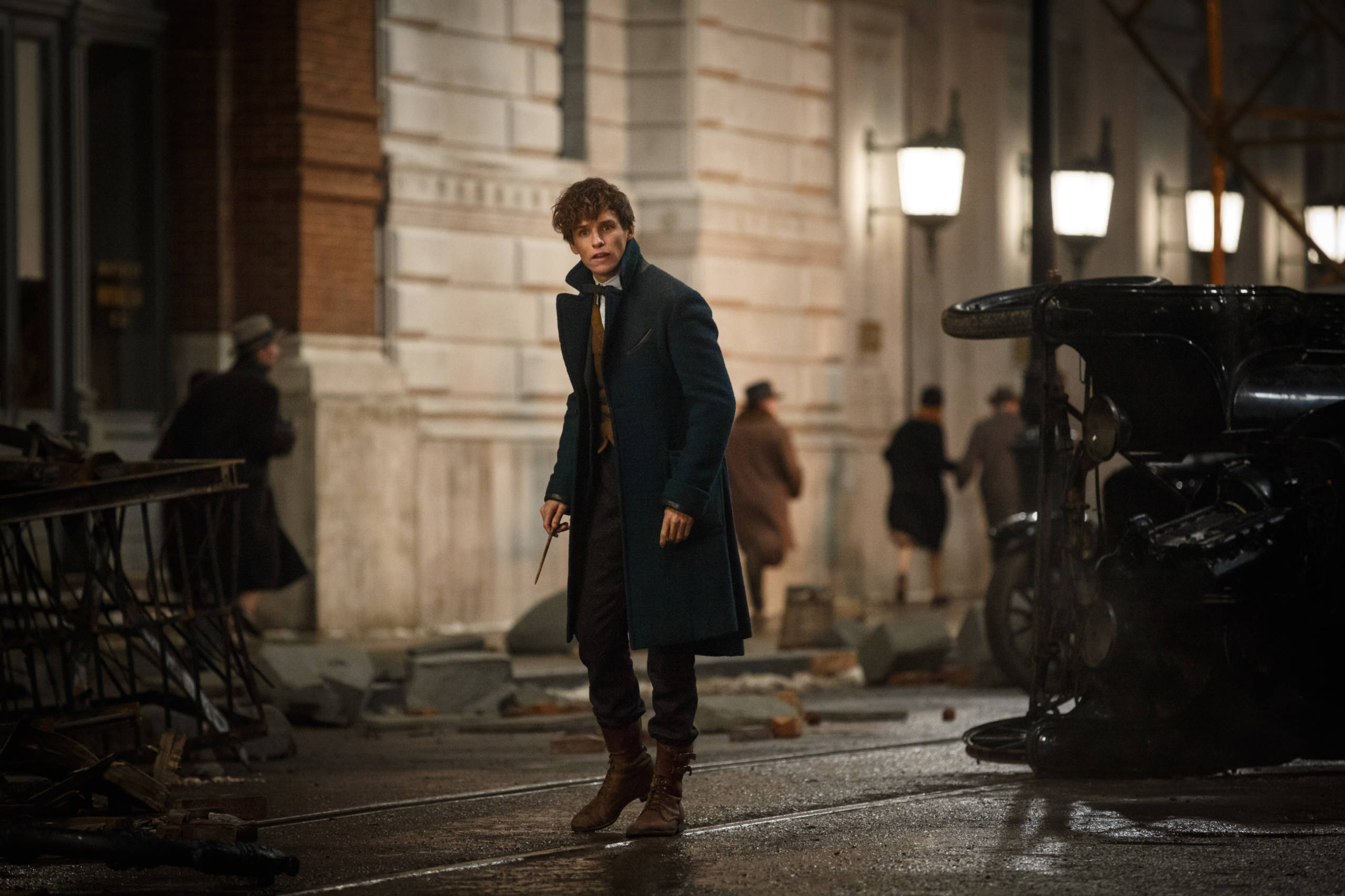 Newt Scamander (Eddie Redmayne) clutches his wand as he advances past upturned cars in Fantastic Beasts and Where to Find Them