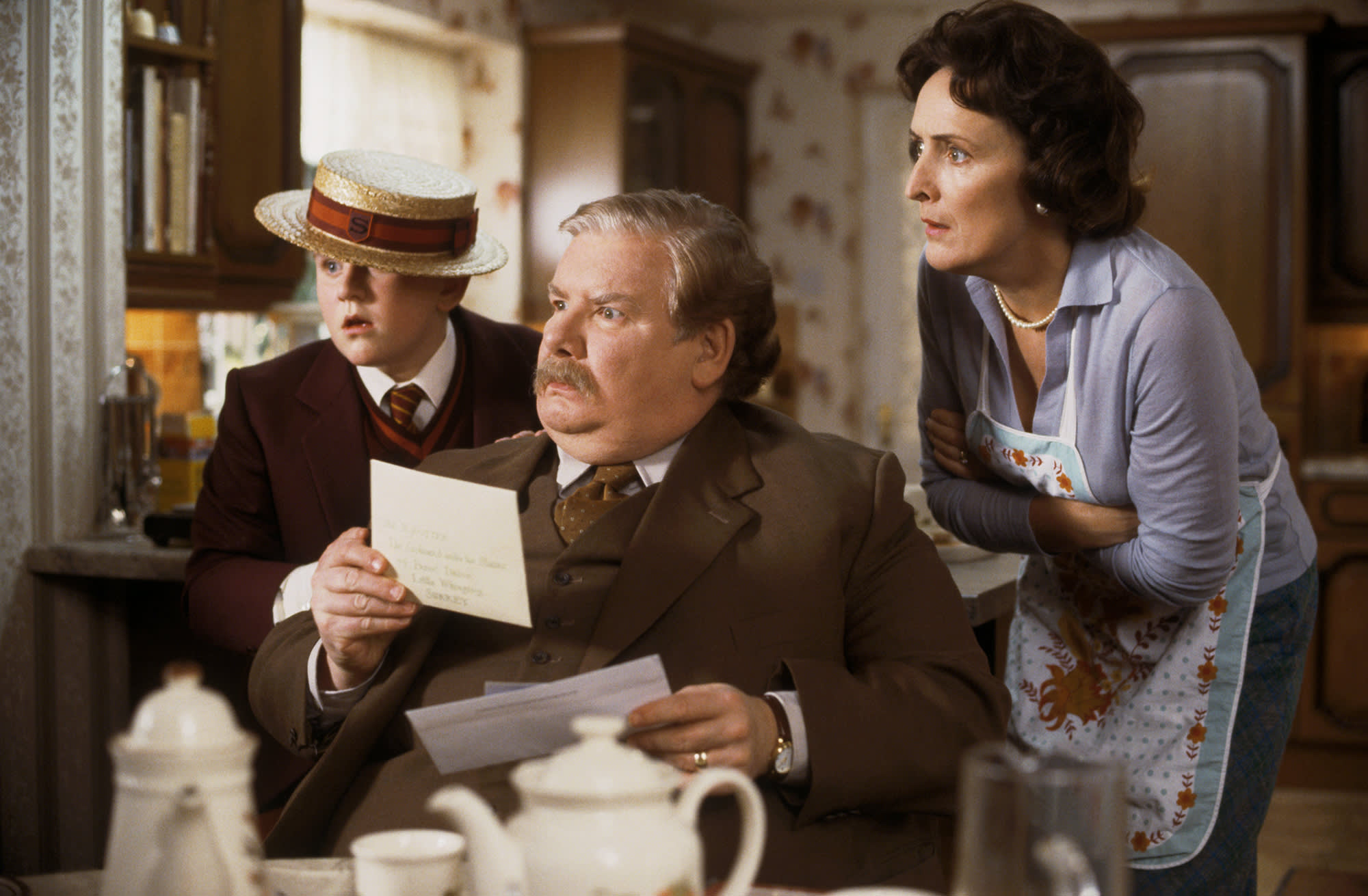 Vernon Dursley sat at the kitchen table holding Harry's Hogwarts letter. Petunia and Dudley are both looking over his shoulder. All three look surprised and concerned.