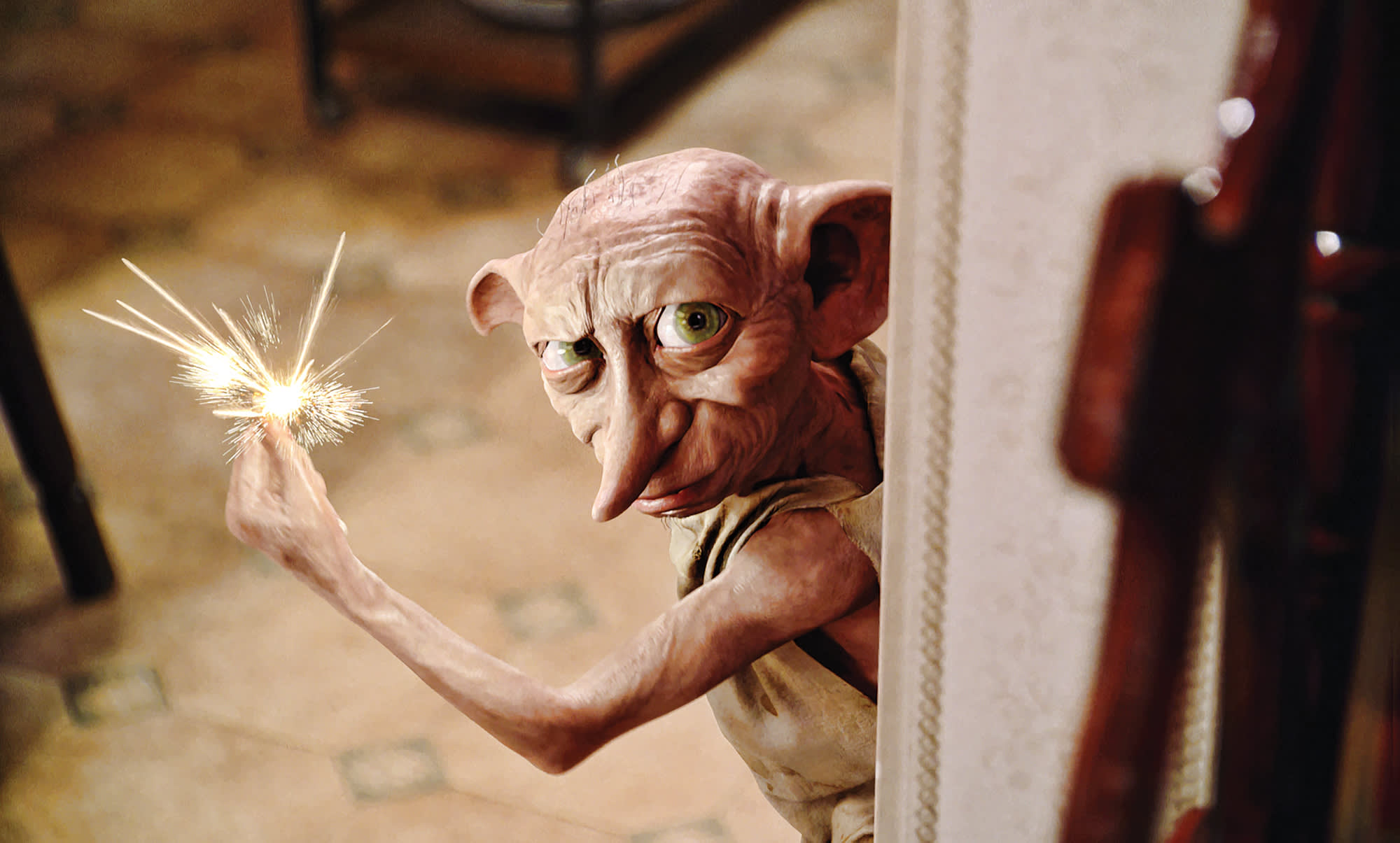 Dobby is looking round a doorway in 4 Privet Drive. He is using magic and snapping his fingers which causes a spark.