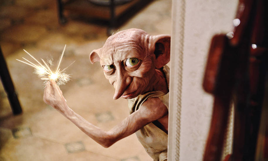 Dobby is looking round a doorway in 4 Privet Drive. He is using magic and snapping his fingers which causes a spark.