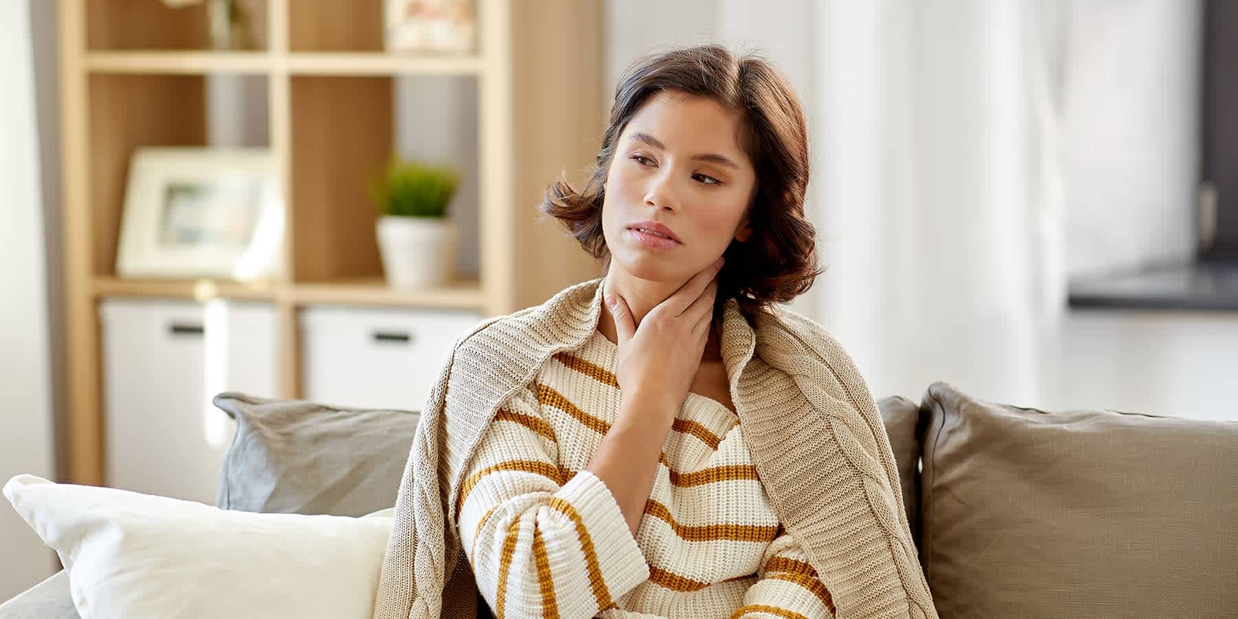 Woman sitting on couch with hand over neck and wondering about hypothyroidism vs. hyperthyroidism