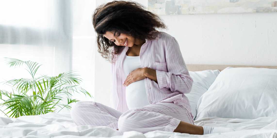 Pregnant woman sitting on bed and smiling while wondering if gestational diabetes is genetic