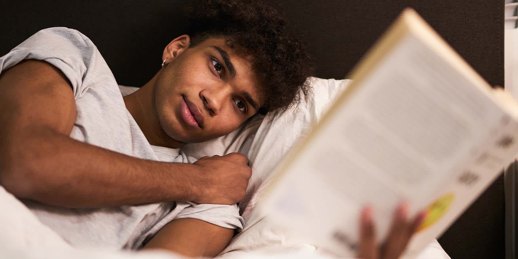 Young man in bed reading book and wondering "Why can't I sleep?"