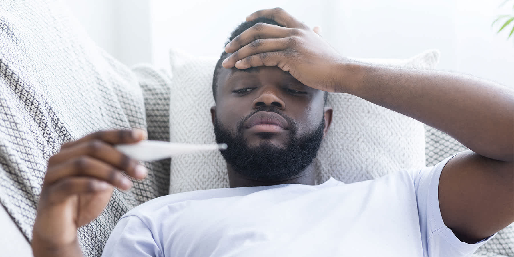 Man with syphilis symptoms lying down while experiencing fever and looking at a thermometer 