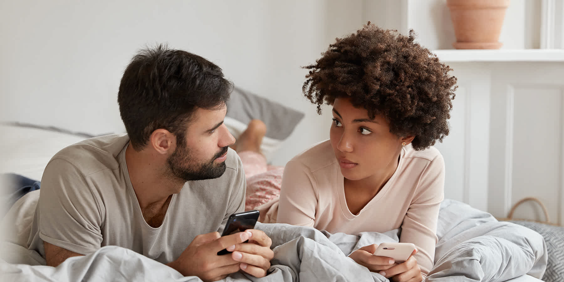 Couple in bed browsing phones and looking at each other while discussing syphilis vs. herpes
