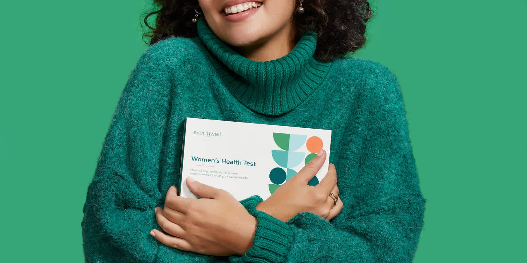 Woman holding an Everlywell Women's Health Test box close to her against a green background