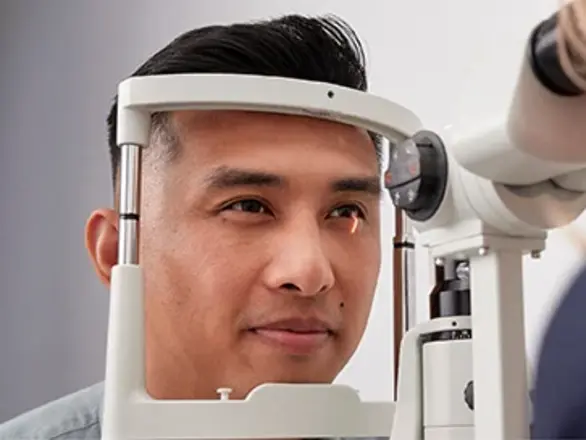 A man undergoes an eye exam, looking into a slit lamp while an eye care professional examines his eye.