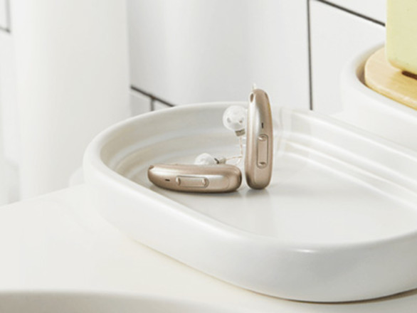 Two beige hearing aids resting on a white ceramic tray in a bathroom setting.