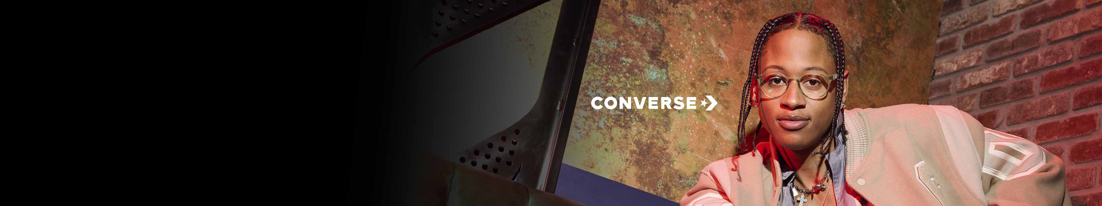 Person with glasses and braids wearing a light jacket sits on a couch; the word "CONVERSE" appears on the textured wall behind them.