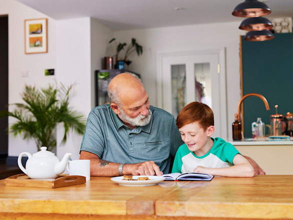 Grandfather reading with grandson at dining table.