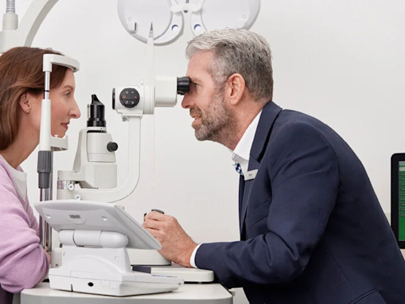 An eye doctor examines a patient with a slit lamp in a clinical setting. Both are seated, and diagnostic equipment is visible on the desk.