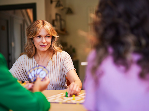 A woman smiles while playing a board game with two others in a cozy, well-lit room.