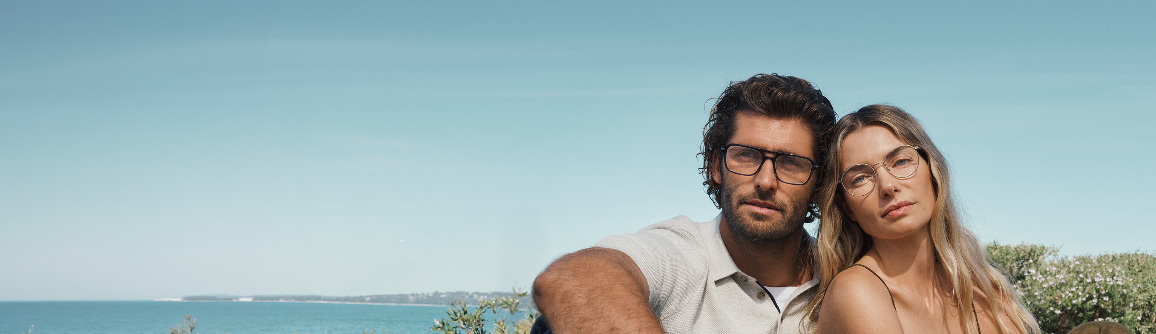 A man and a woman wearing glasses sit closely together outdoors under a clear blue sky, looking directly at the camera.