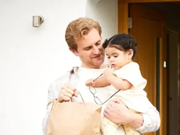 A man holding a young child and a brown paper bag stands outside a house near an open door.