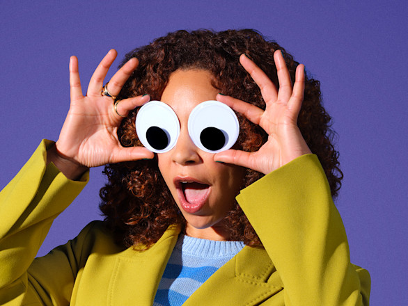 A person in a yellow jacket holds large googly eyes over their face against a purple background, with an open-mouthed expression.