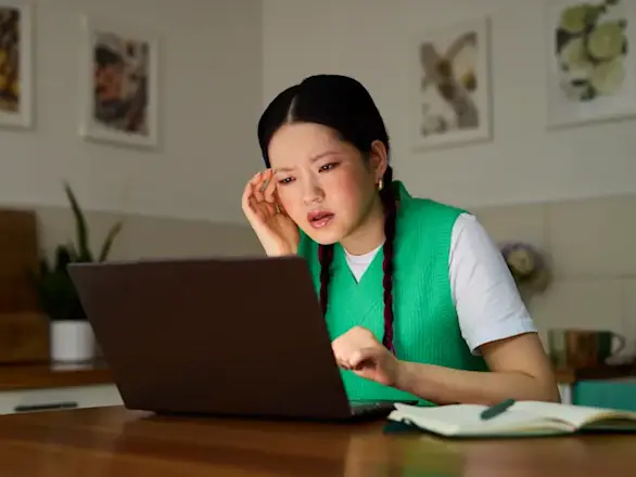 A woman in a green vest sits at a kitchen table, looking confused or concerned while using a laptop, with a notebook nearby.