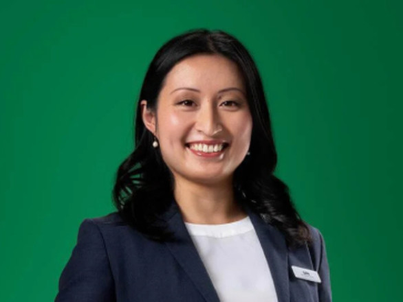A woman with long dark hair, wearing a navy blazer and white shirt, smiles in front of a plain green background.