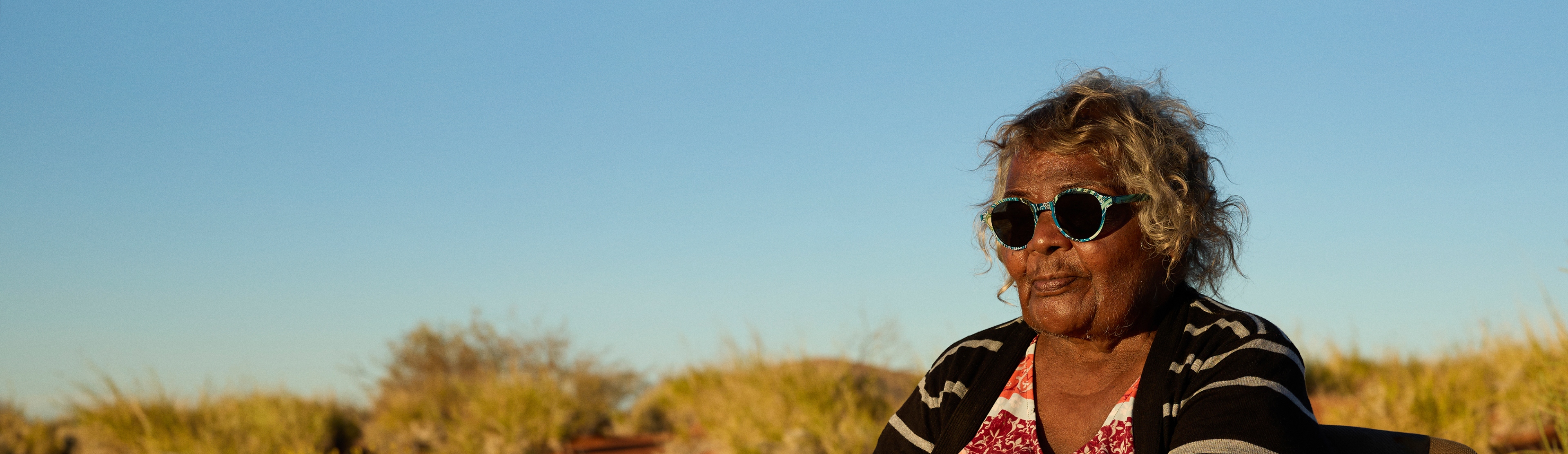 An older Indigenous woman wears Fred Hollows Limited Edition sunglasses, sits outdoors with a clear blue sky in the background.