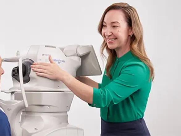 A optometrist operates an eye examination machine while an older man sits in front of it.